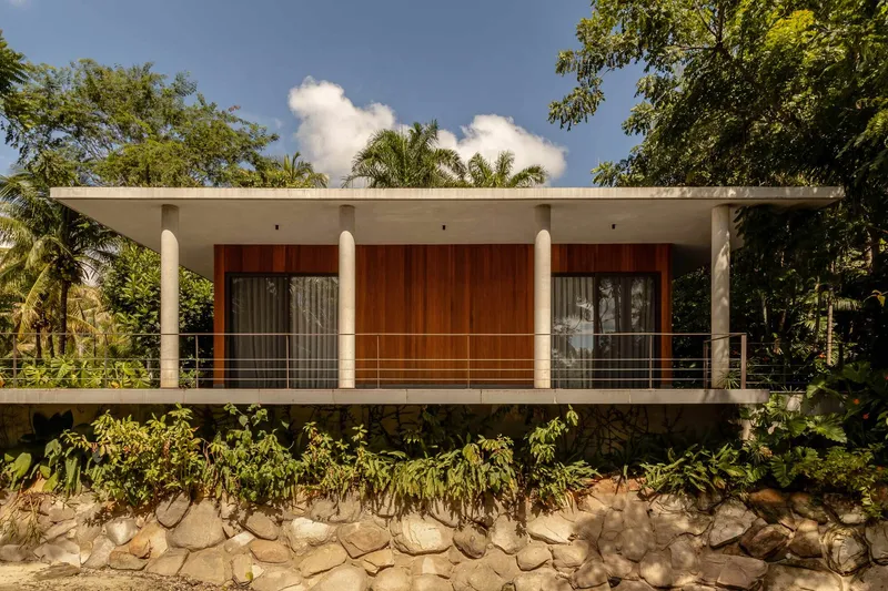 Elevated concrete volume with timber-clad facade and cable railing above a stone-retained embankment surrounded by palms