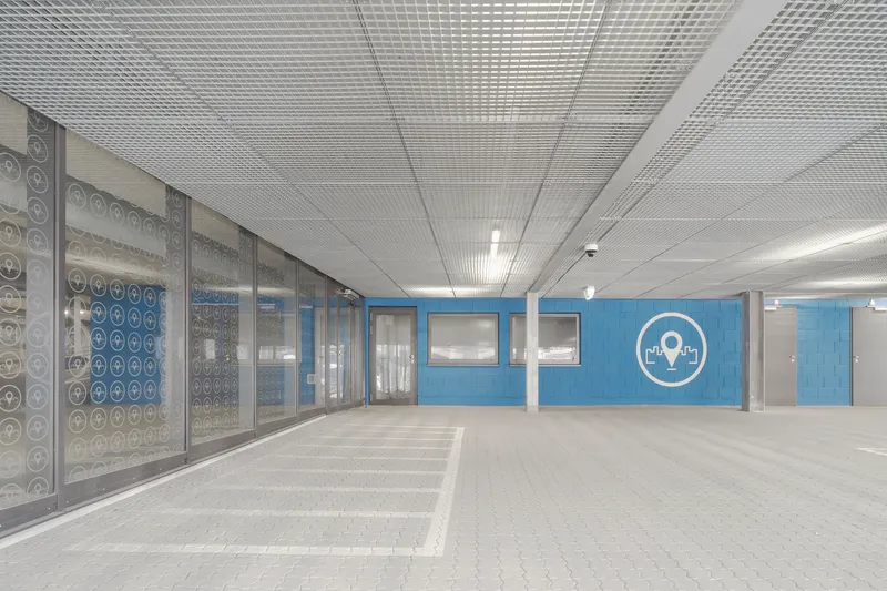 Interior parking level with perforated glass panels and blue painted wall beneath a metal mesh ceiling