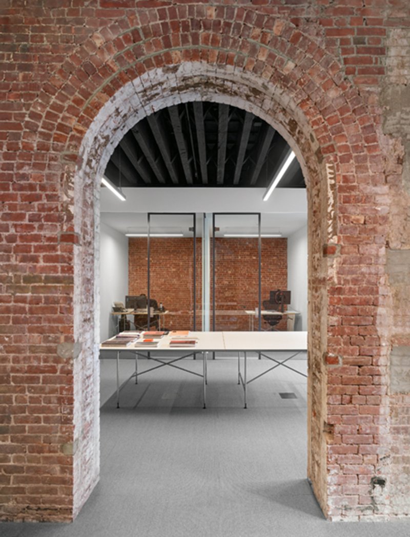 View through exposed brick archway into gallery space with display table and suspended brick panels
