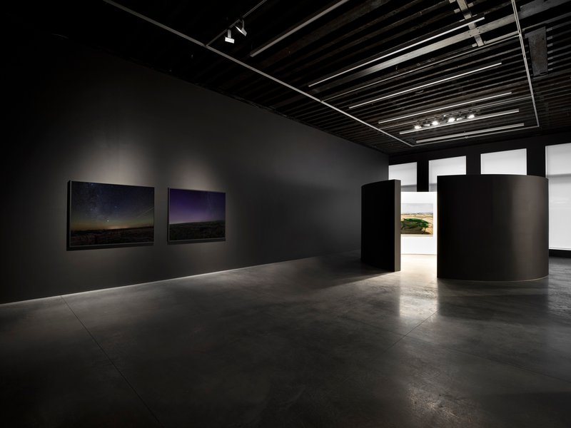 Dark gallery space with charcoal walls, exposed black ceiling joists and cylindrical partition element