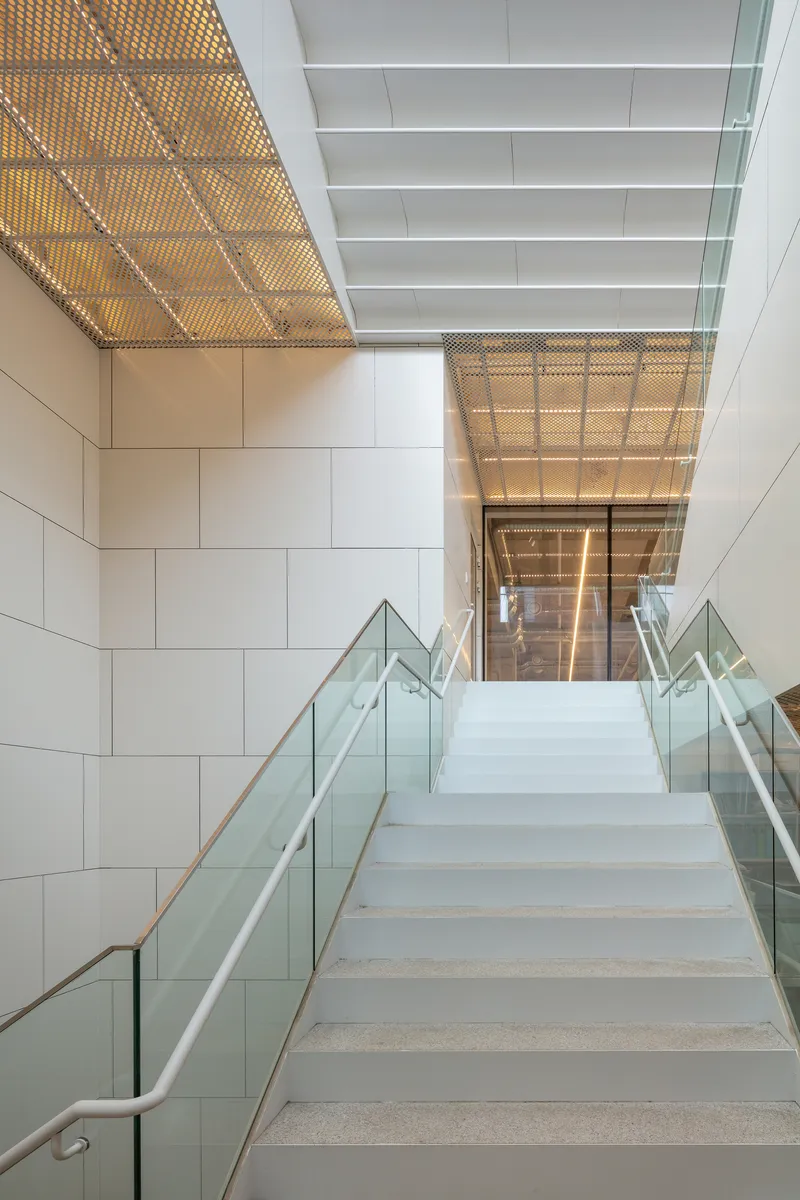 Stairwell with perforated metal ceiling panels and glass balustrades lit by warm afternoon light