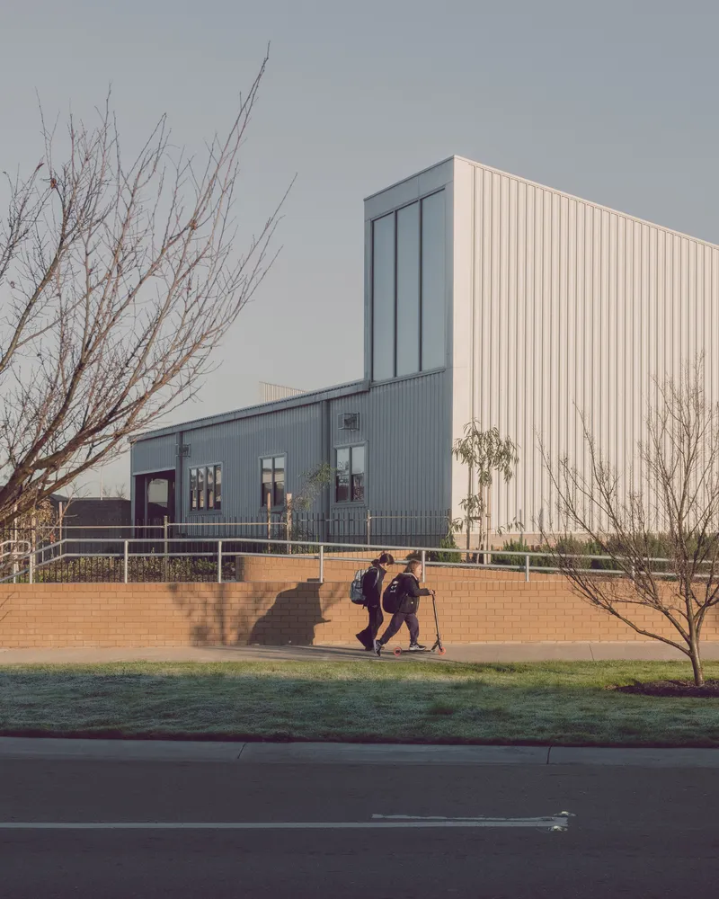 Vertical tower clad in cream corrugated metal rising above horizontal classroom wing with two students walking past