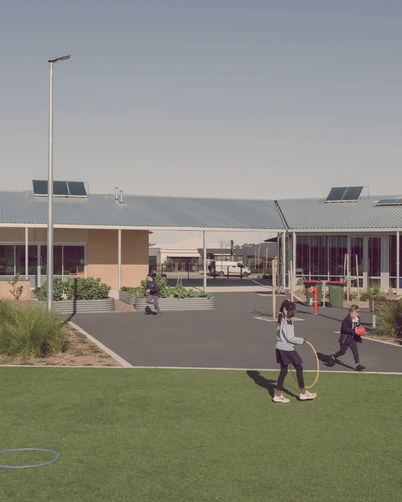 Courtyard view with children playing on synthetic turf and asphalt paths between low-slung buildings with corrugated metal roofs and solar panels