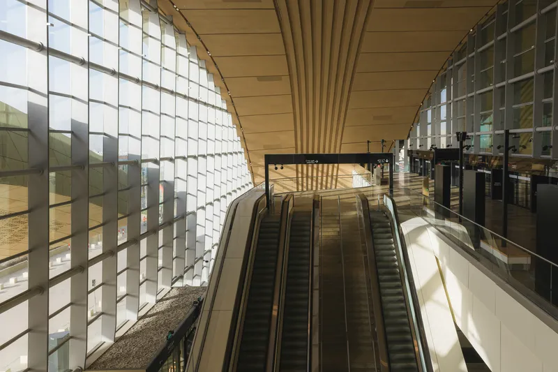 Escalators descending through a hall with gridded glass walls and a radiating timber ceiling