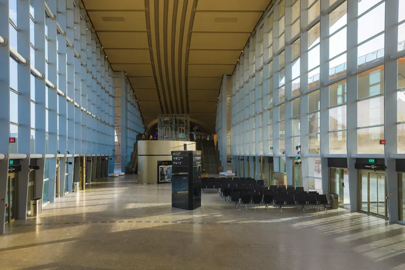 Long hall with gridded glass facades and rows of black seating under a ribbed timber ceiling