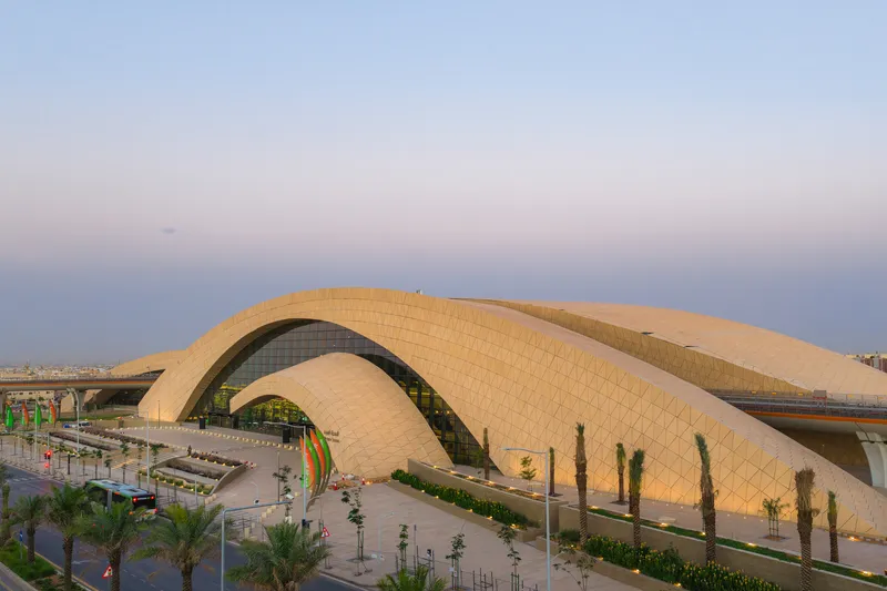 Double-arched roof clad in diamond-pattern tiles spanning above glass-walled entrance at dusk