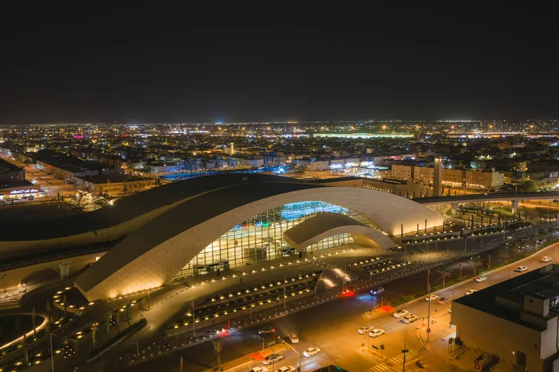 Night aerial showing the illuminated arched roof above the roadway with city lights in the distance