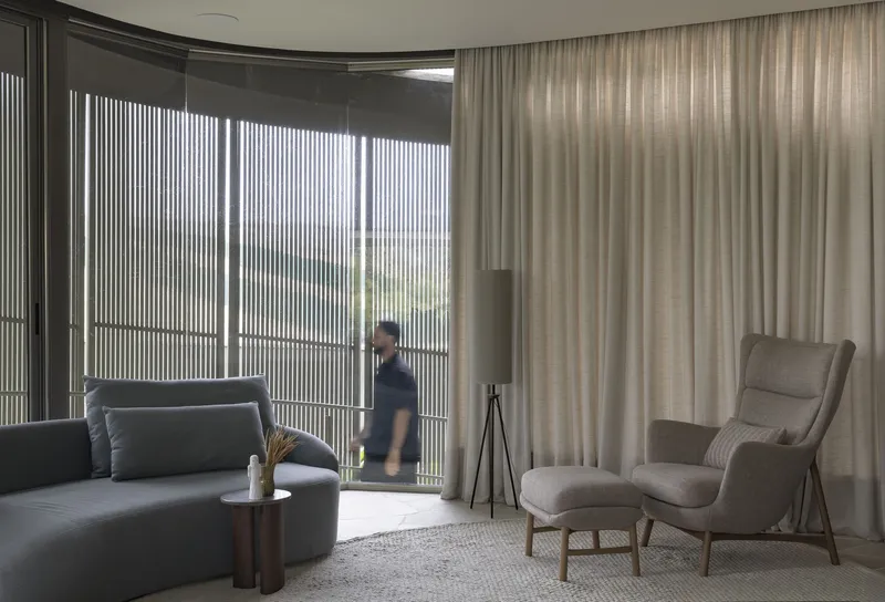 Curved living room with vertical metal screen doors and sheer curtains filtering daylight with a person passing through