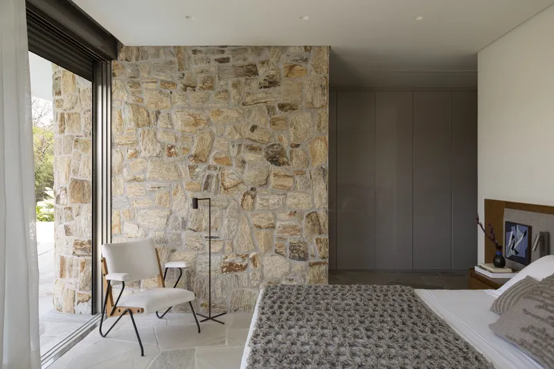 Bedroom with rough-cut stone wall adjacent to floor-to-ceiling glazing and a chair beside the window