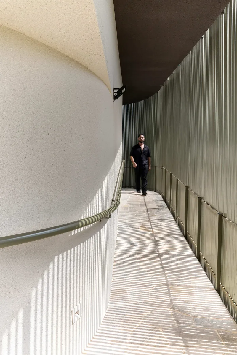 Corridor with ribbed metal wall casting striped shadows beside a curved white plaster surface with a person walking