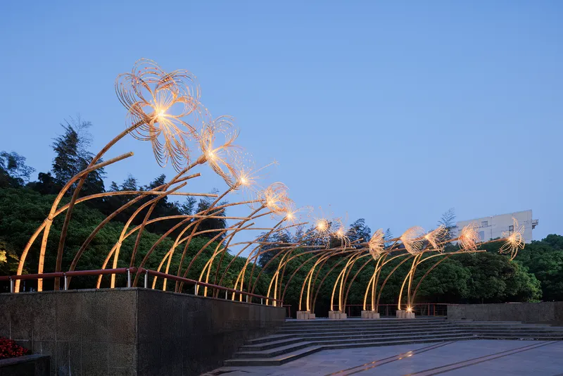 Illuminated steel arch installation with wire blossom forms spanning a public plaza at dusk