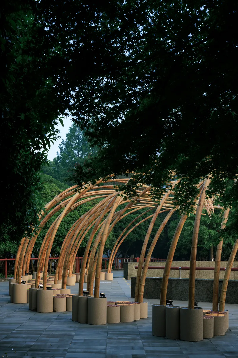 Arched bamboo structure anchored in cylindrical concrete bases on a paved plaza framed by trees at dusk