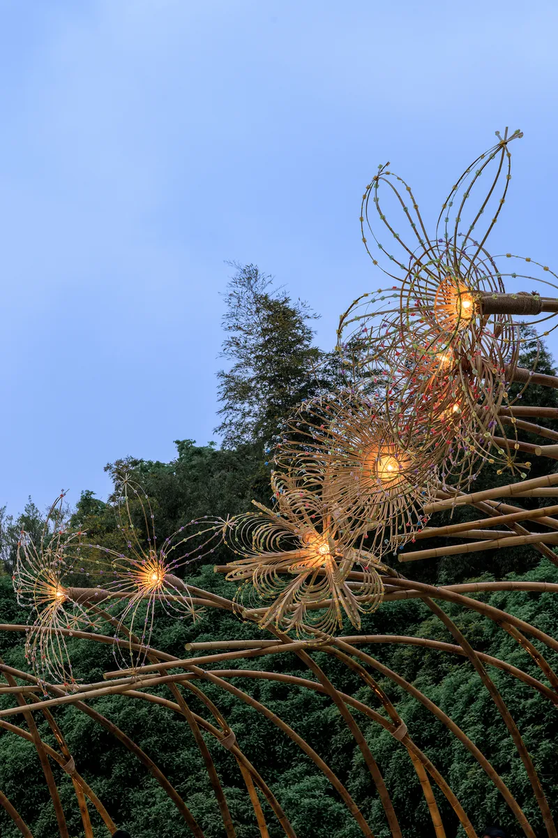 Bamboo sculpture with integrated lighting illuminating wire flower forms at dusk among surrounding trees