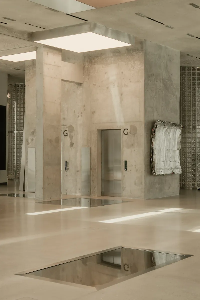 Two elevator doors flanked by concrete walls with rectangular floor mirror reflecting light