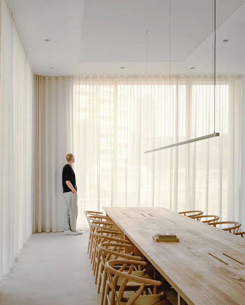 Dining room with long timber table and linear pendant light as figure stands by sheer curtains