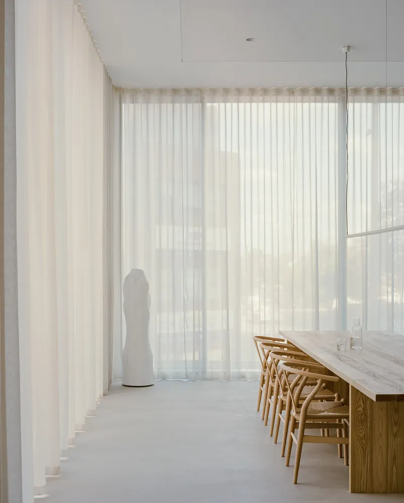 Corner of dining space with white sculptural form beside floor-to-ceiling sheer curtains in daylight