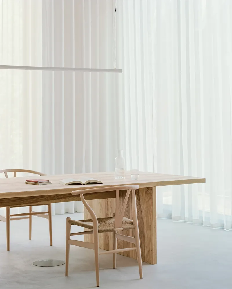 Close view of timber dining table with open book and linear pendant against backlit sheer curtains