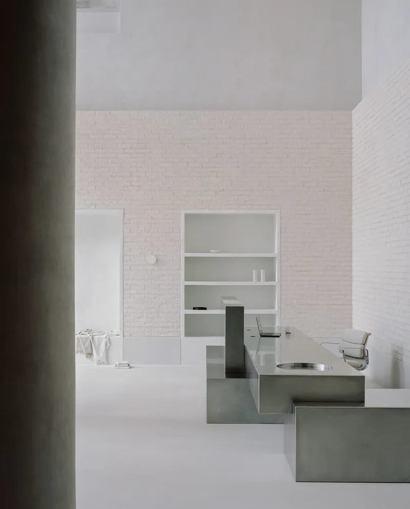 Minimalist workspace with white painted brick walls, built-in shelving, and a stainless steel desk under soft natural light