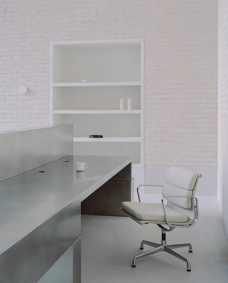 Close-up of a polished steel desk with recessed shelving and a leather office chair against white brick