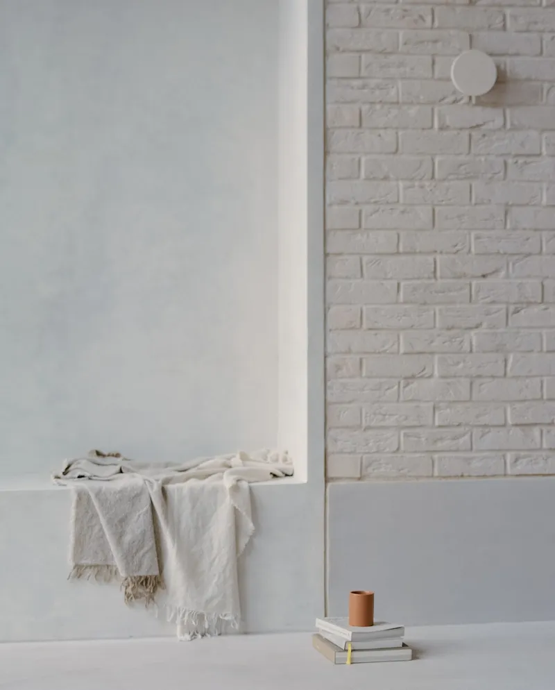 White brick wall with circular sconce above a ledge holding folded linens and stacked books