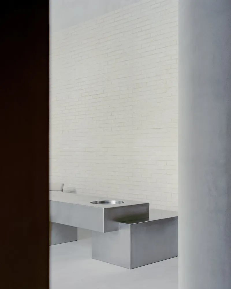 Stainless steel sink with circular basin inset into floating counter against white brick wall in shadow