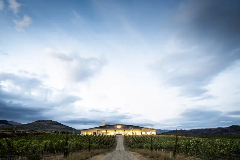 Night view of illuminated low-slung building with central tower set among vineyard rows under dramatic sky
