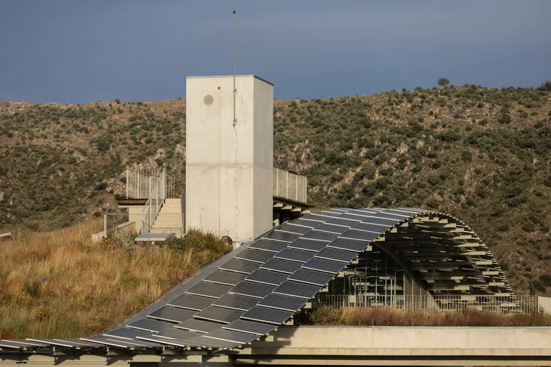 Curved solar panel canopy beside white concrete tower with hillside and scrubland in background