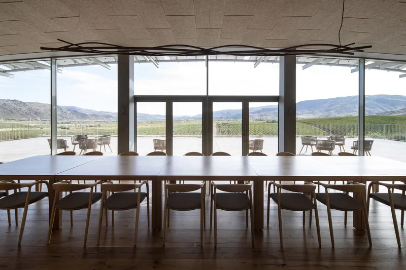 Interior dining room with long table and glazed walls overlooking vineyard rows and distant mountains