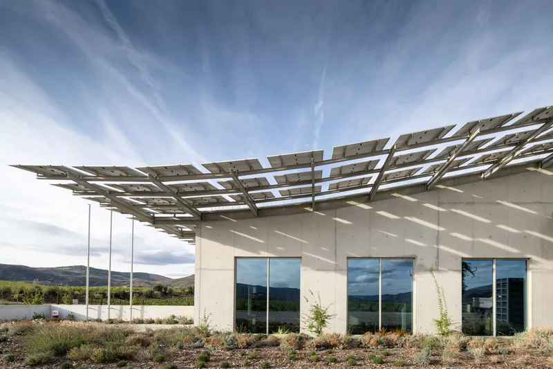 Corner view of white facade with photovoltaic canopy casting dappled shadows on planted surroundings