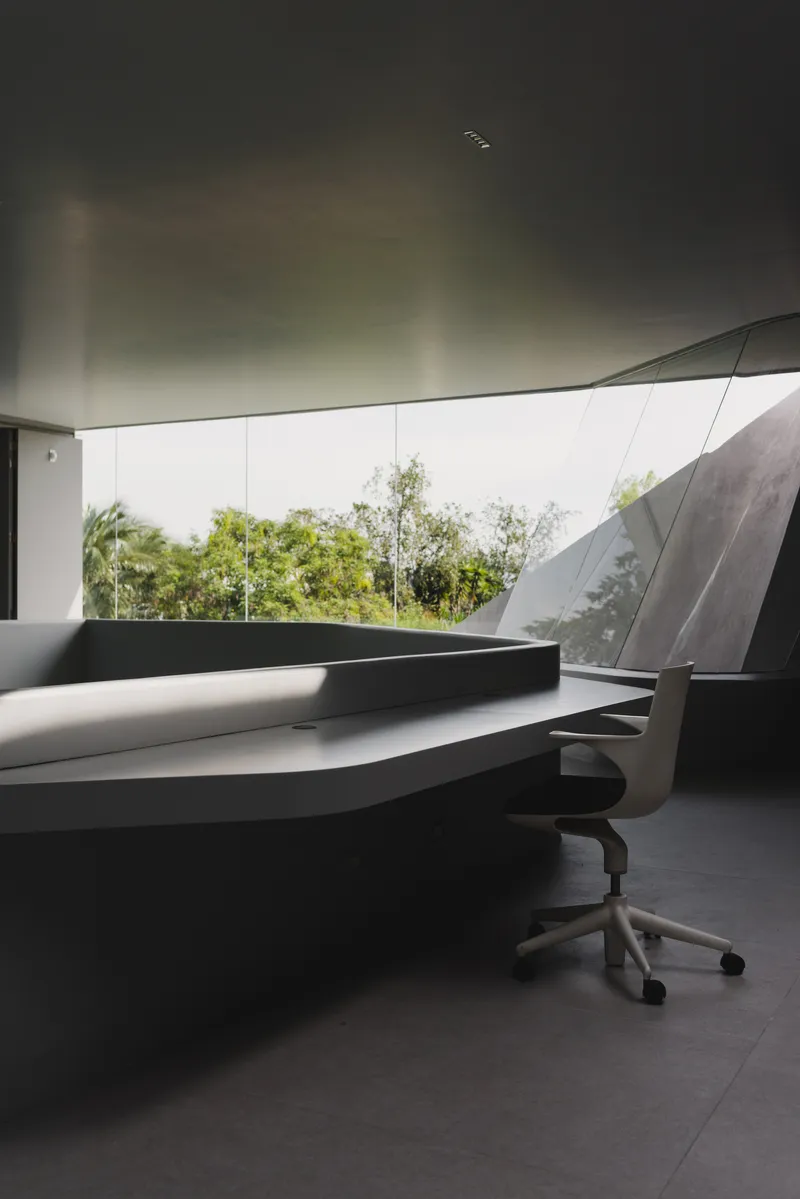 Dark desk workspace beneath a curved soffit with frameless glass opening to trees beyond