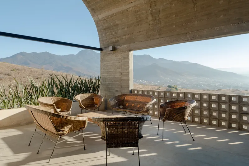 Covered terrace with woven rattan seating beneath board-formed concrete vault overlooking distant mountains