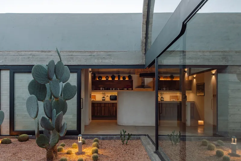 Glass-walled interior kitchen and dining space overlooking a gravel courtyard with cacti illuminated at dusk