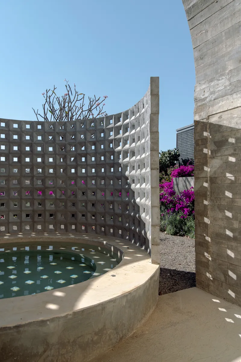 Curved perforated concrete wall enclosing a shallow plunge pool with glimpses of magenta flowers beyond