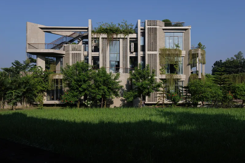 Side facade with latticed concrete panels and cascading vegetation viewed across a paddy field at dusk