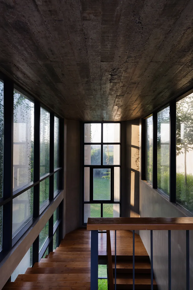 Interior bridge with board-formed concrete ceiling and floor-to-ceiling windows overlooking a courtyard garden