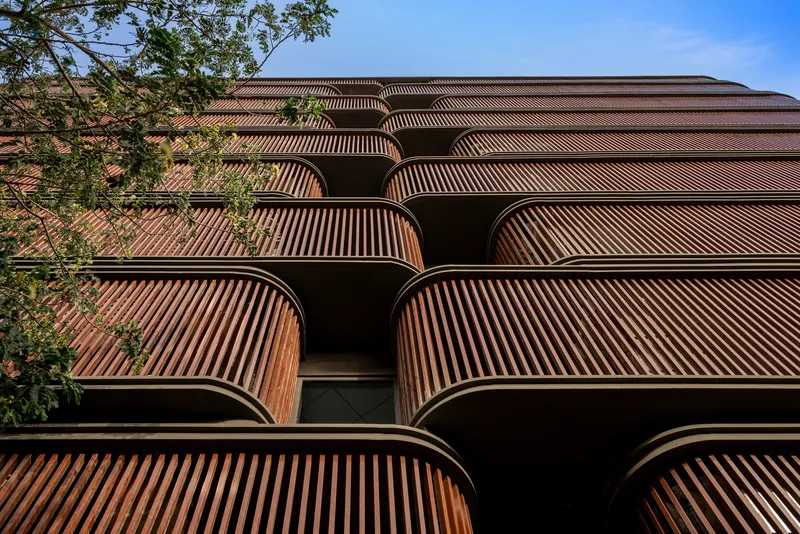 Stacked facade of curved timber louvered balconies framed by green foliage and blue sky