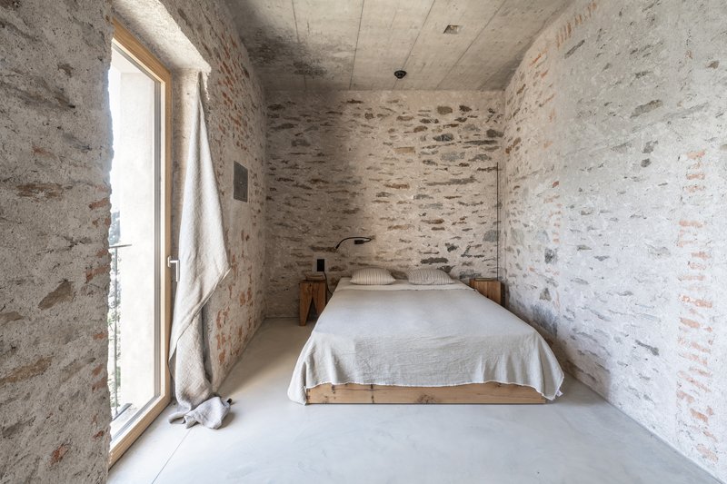 Bedroom with whitewashed stone walls, concrete ceiling, and timber bed frame beside a floor-to-ceiling window