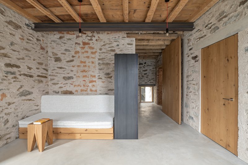 Living space with exposed timber roof beams, steel column, and whitewashed stone walls under natural light