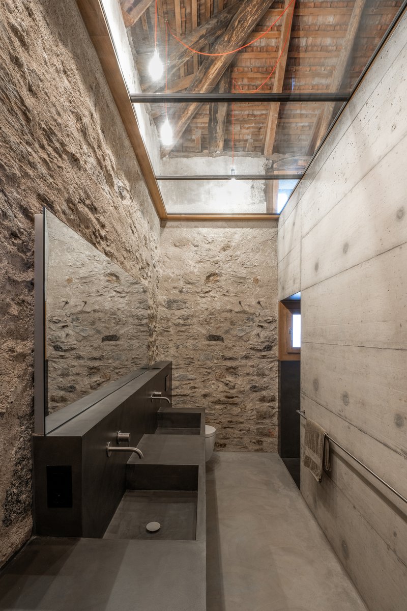 Bathroom with overhead skylight, exposed timber roof structure, and concrete vanity counter against stone walls