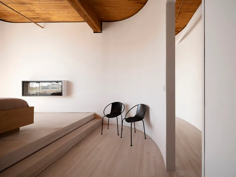 Curved platform with two black chairs beside a low horizontal window under vaulted timber ceiling
