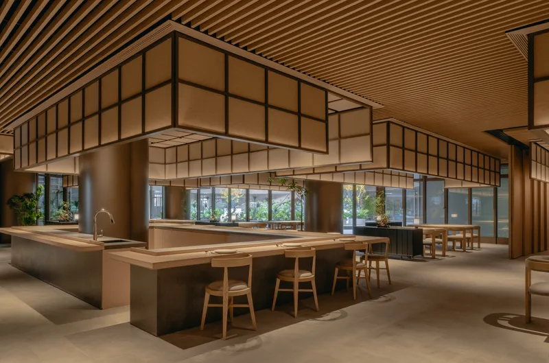 Long interior view showing communal timber table, slatted ceiling and glazed storefront opening to exterior greenery