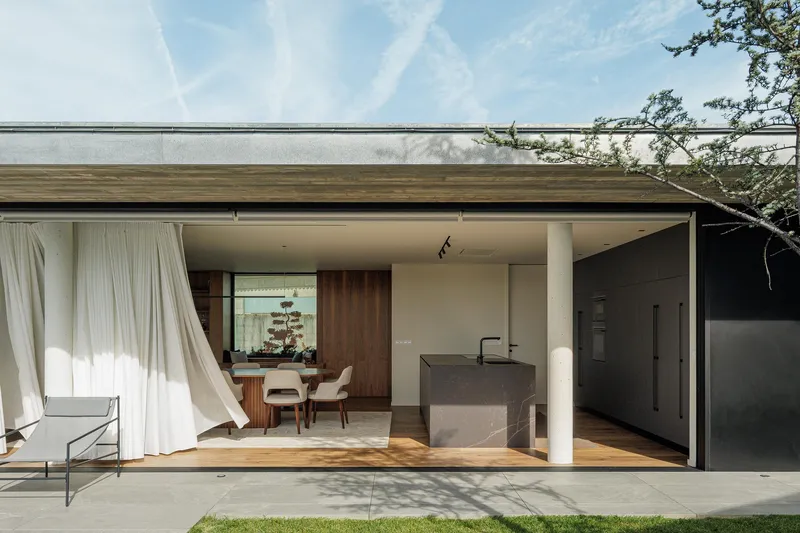 Covered outdoor terrace with flowing curtains opening to kitchen island and timber-clad dining area beyond
