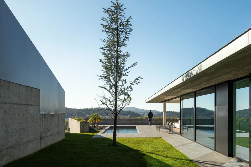 Courtyard view with concrete and glass volumes framing a slender tree and swimming pool under a clear sky