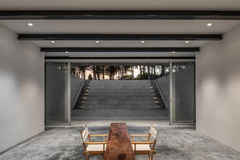 Interior room framing a concrete staircase ascending to trees at dusk through full-height glazing