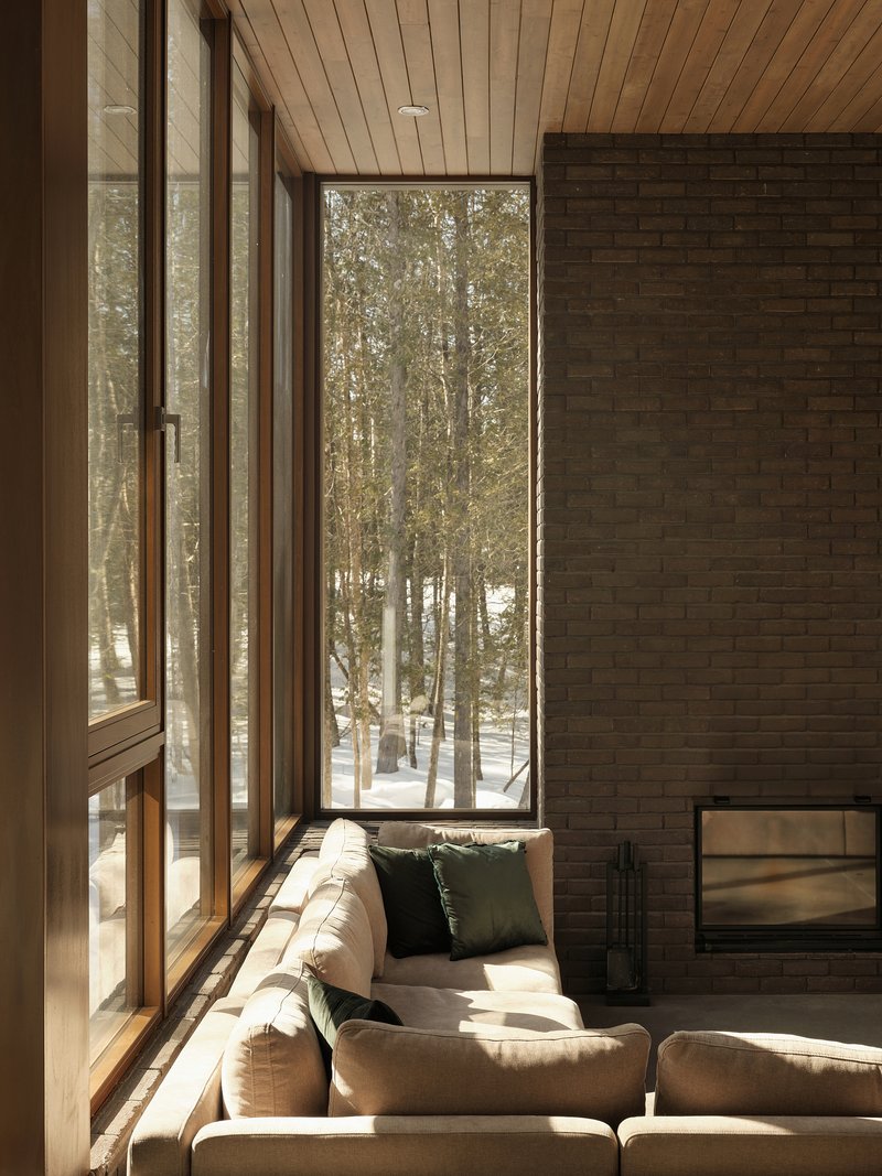 Living room with floor-to-ceiling windows, brick fireplace wall and wood plank ceiling overlooking winter trees