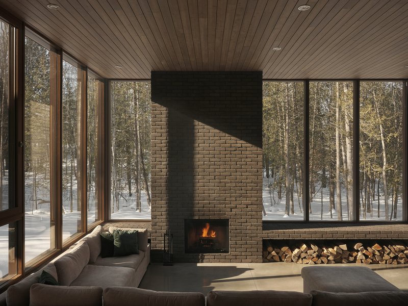 Brick fireplace with active fire flanked by glass walls and built-in firewood storage in winter sunlight