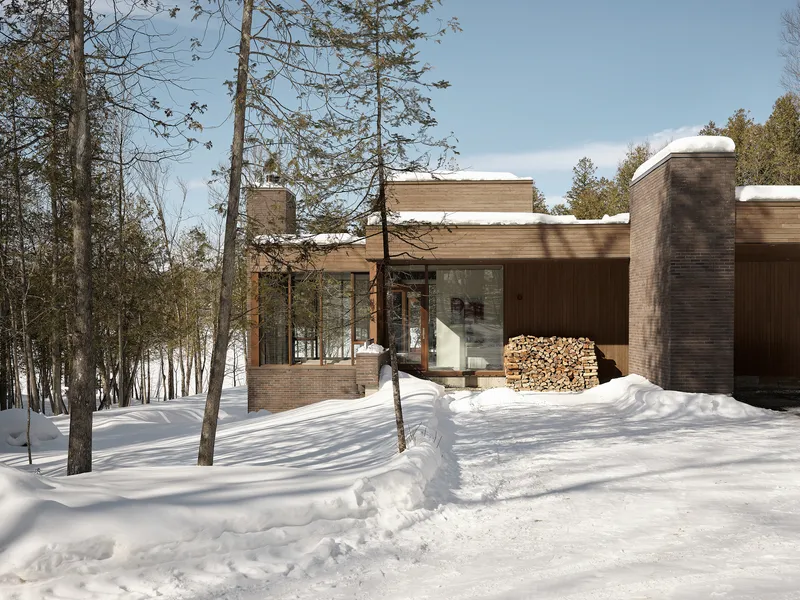 Brick facade with flat roofs covered in snow and stacked firewood beside glass doors among bare trees