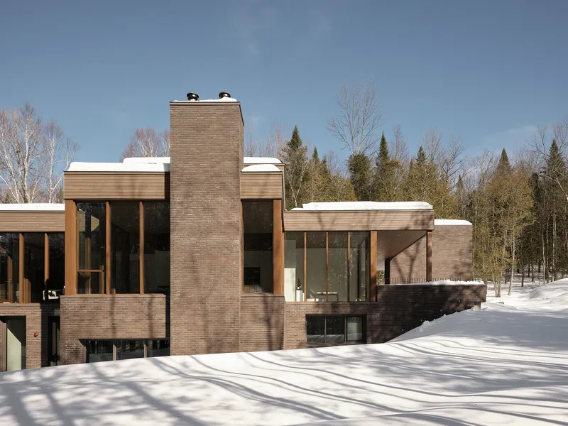 Multi-level brick facade with chimney and glass walls nestled in a snowy forest clearing