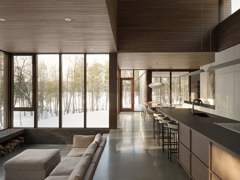 Kitchen island with white cabinetry and horizontal wood-planked ceiling looking toward winter landscape through glass walls