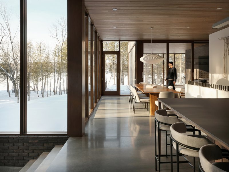 Dining and kitchen space with floor-to-ceiling glazing overlooking snow-covered forest and a figure standing
